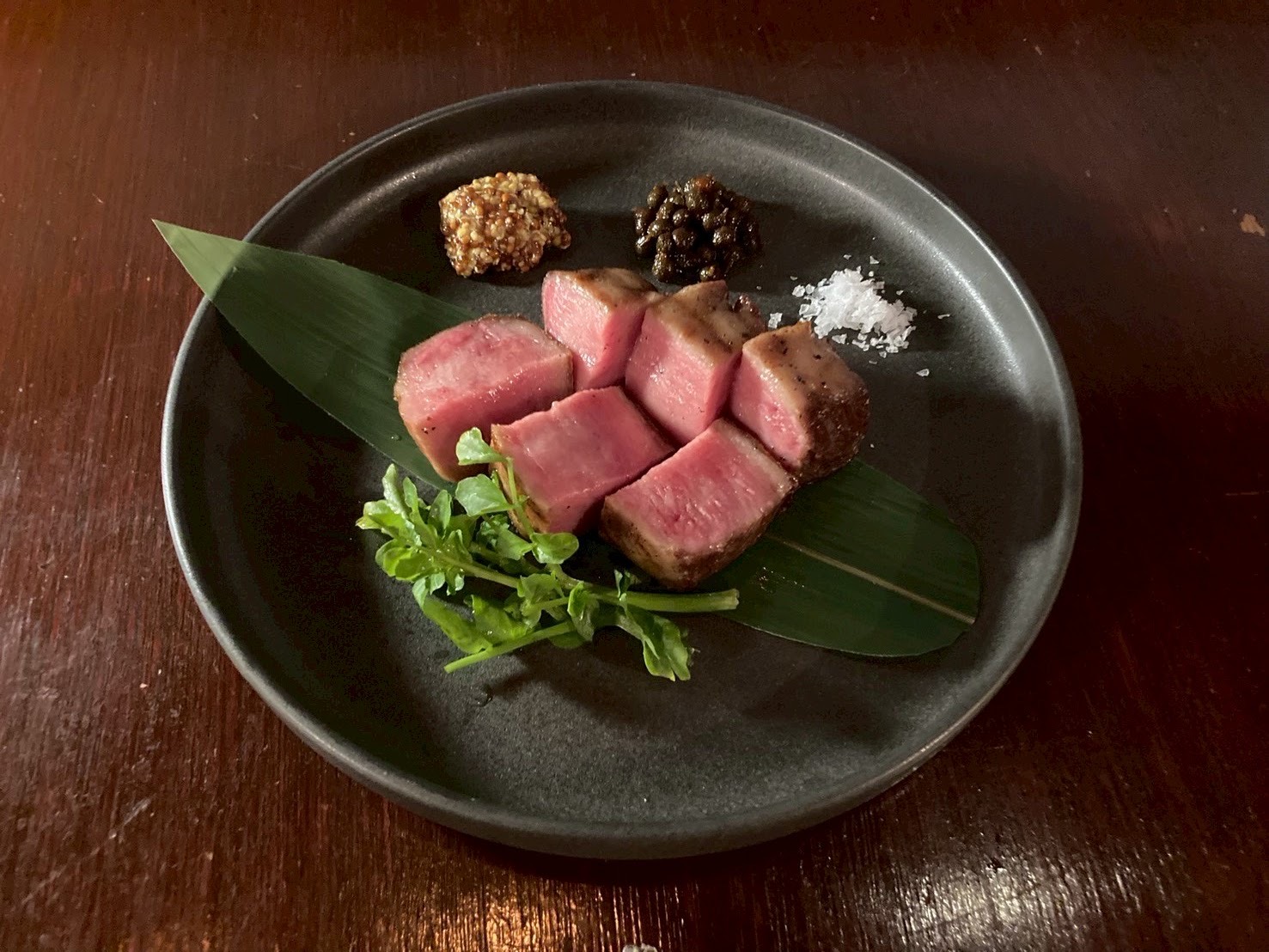 黒い皿に笹の葉を敷き、焼き目をつけた厚切りの肉。粗塩と二種の薬味、クレソンを添えて。