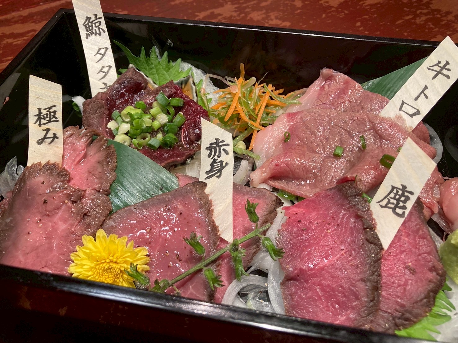 黒い重箱に盛られた肉の盛り合わせ。「鯨タタキ」「極み」「赤身」「牛トロ」「鹿」の札、黄色い菊と刻みねぎ。